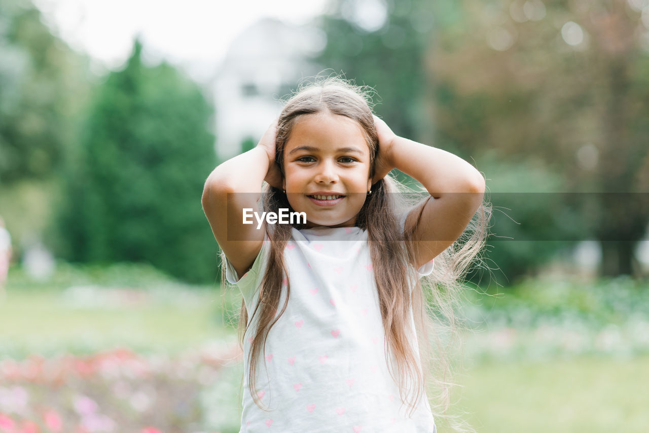 Little girl holds her hands on her head on a walk in the park in summer and smiles