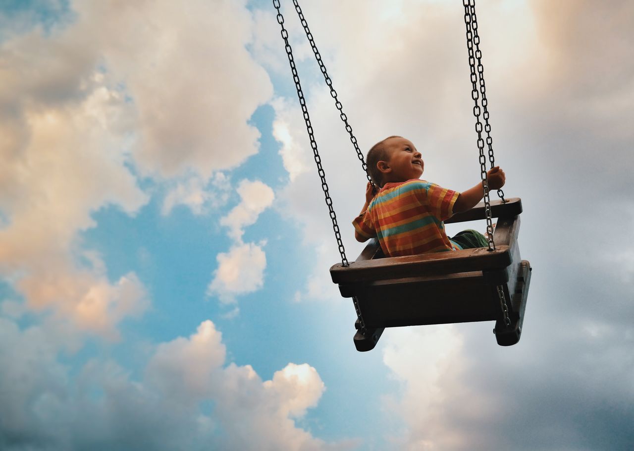 Rear view of smiling boy on swing at playground against sky