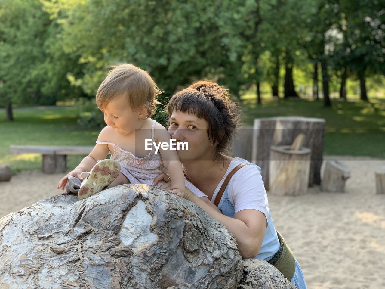 Dark-haired woman standing at big atom inside park and leaning a stone while hugging her chils