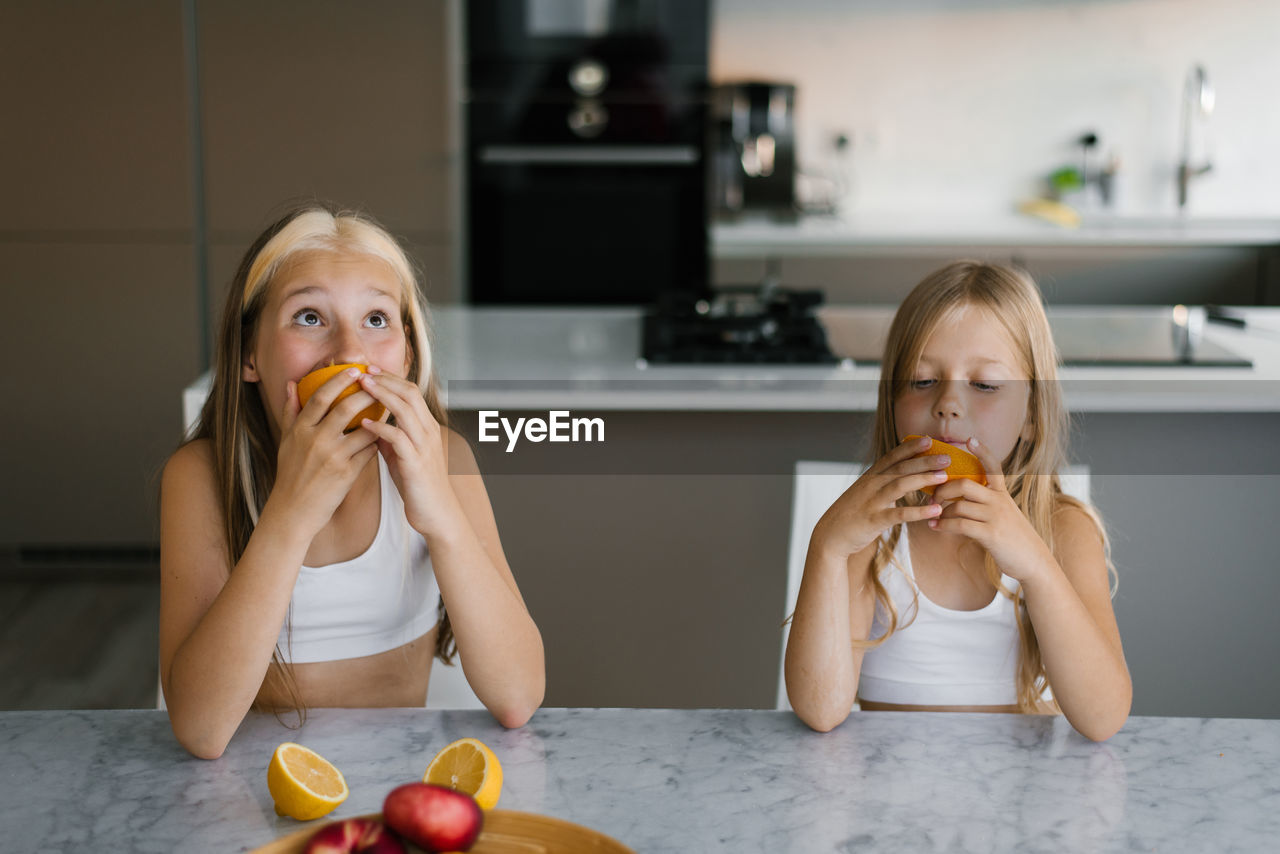 Funny girls children sisters are sitting at the table in the kitchen and eating oranges