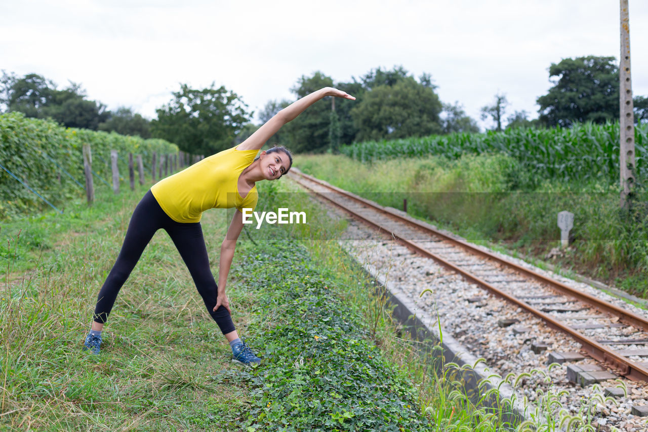 Positive young hispanic sportswoman warming up near railroad tracks in countryside