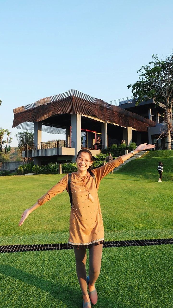 Portrait of young woman with arms outstretched standing on grassy lawn against building