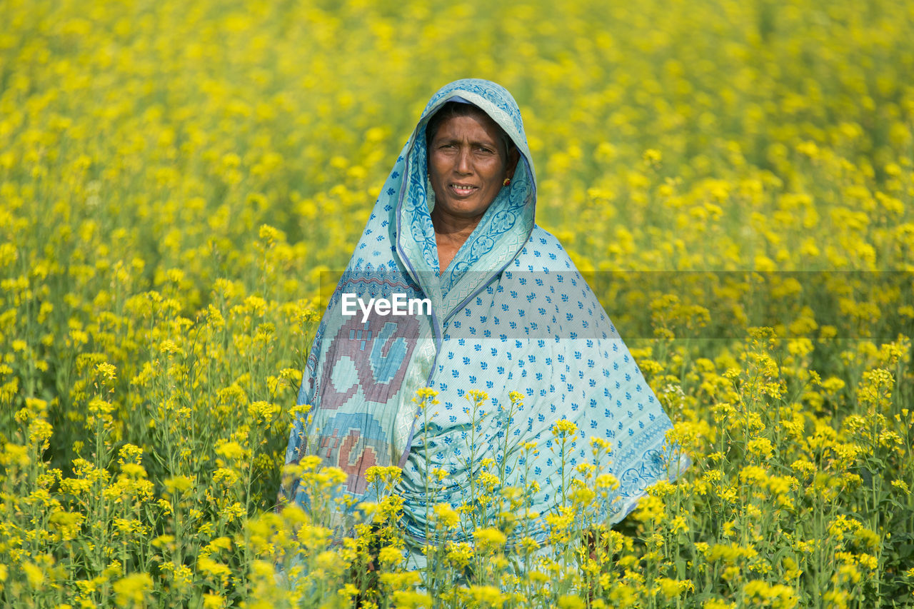 PORTRAIT OF SMILING YOUNG WOMAN STANDING ON YELLOW FLOWER FIELD