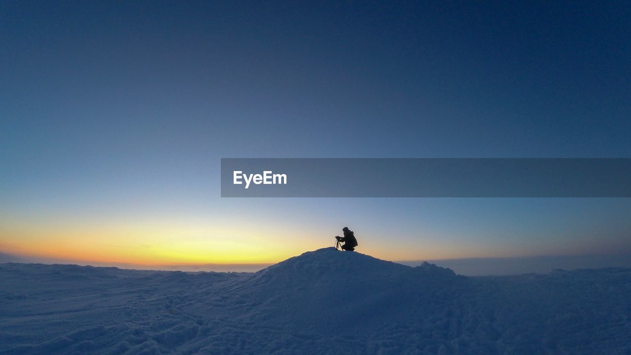 Man crouching on snow covered landscape against clear sky during sunset