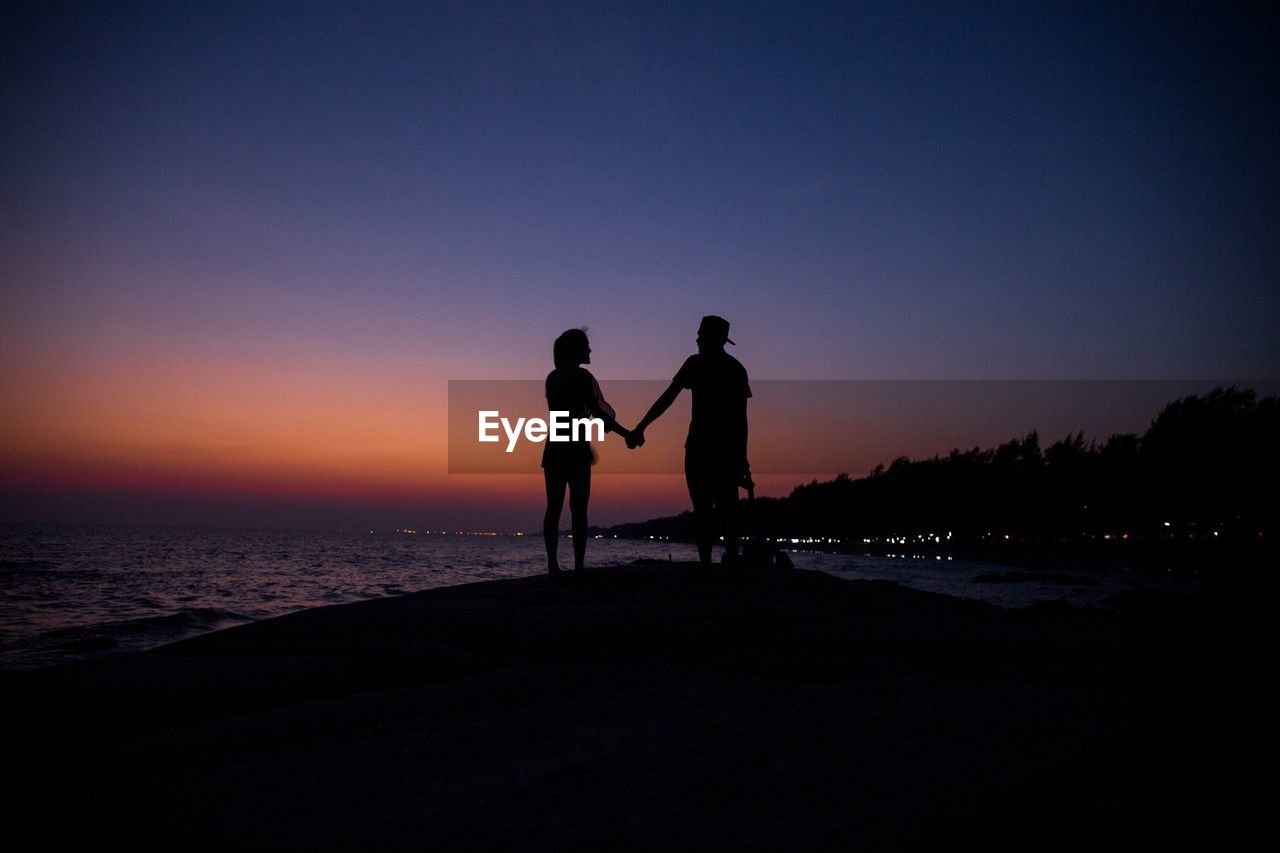 Silhouette of couple holding hands while standing at beach against sky during sunset