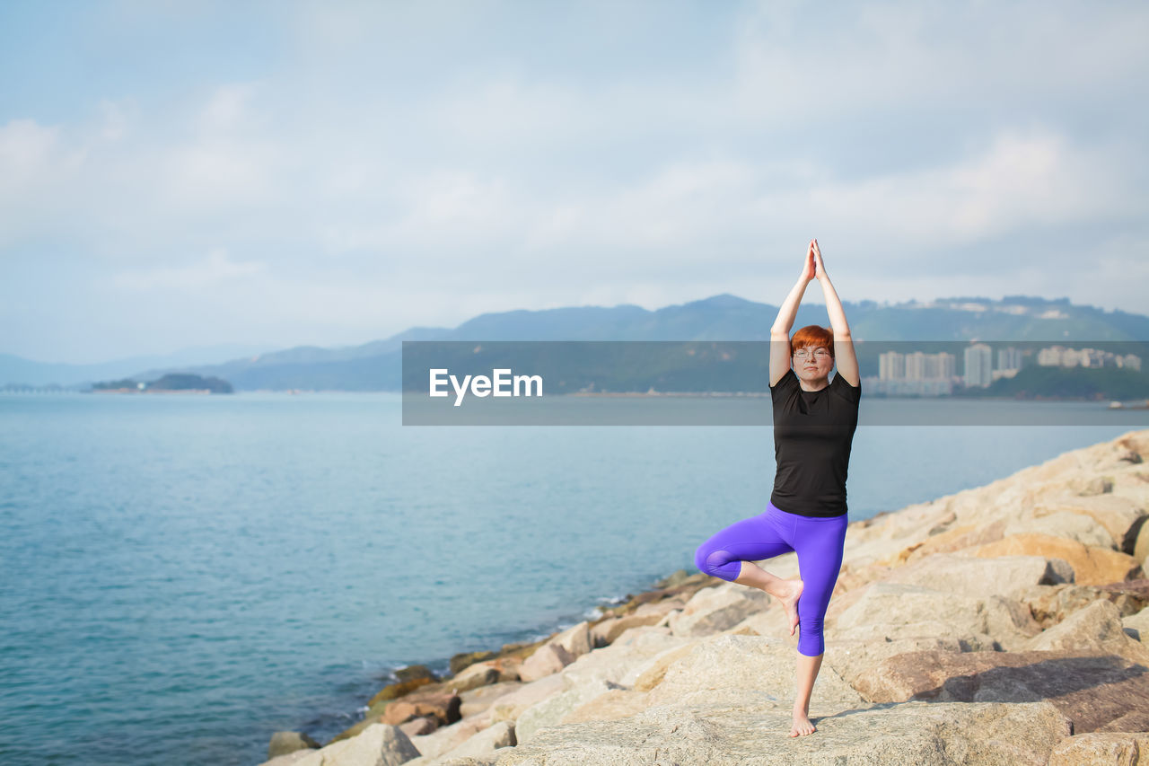 Full length of woman practicing yoga on rock by sea against sky