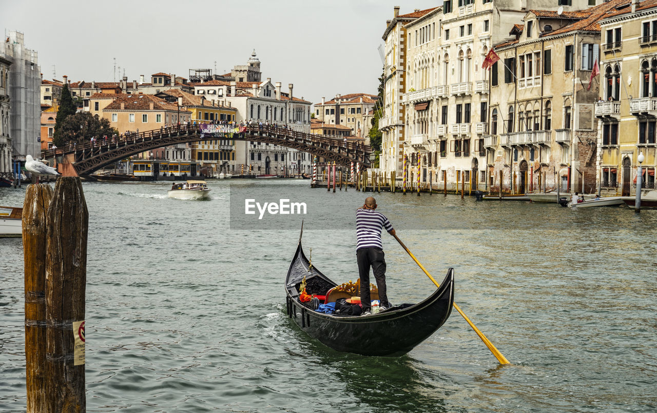 gondola, nautical vessel, transportation, mode of transportation, canal, water, gondolier, architecture, travel destinations, travel, tourism, boat, building exterior, vehicle, built structure, city, nature, bridge, watercraft, tourist, trip, vacation, boating, holiday, romance, rowing, love, waterway, outdoors, oar, men, journey, waterfront, day, adult, on the move, occupation, sky, lagoon, sea, group of people, wooden post, history, building