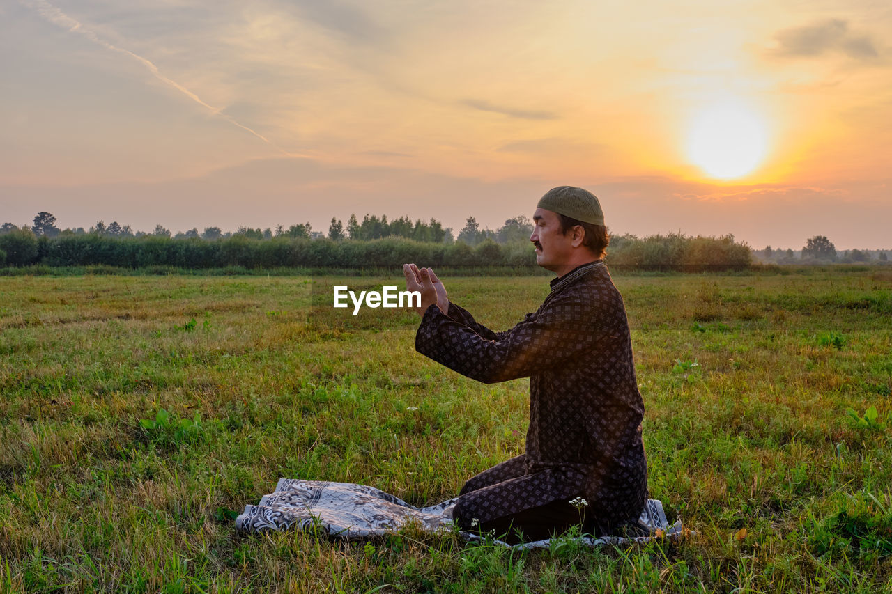 A muslim senior man wearing a skullcap and traditional clothes prays at sunset in a field