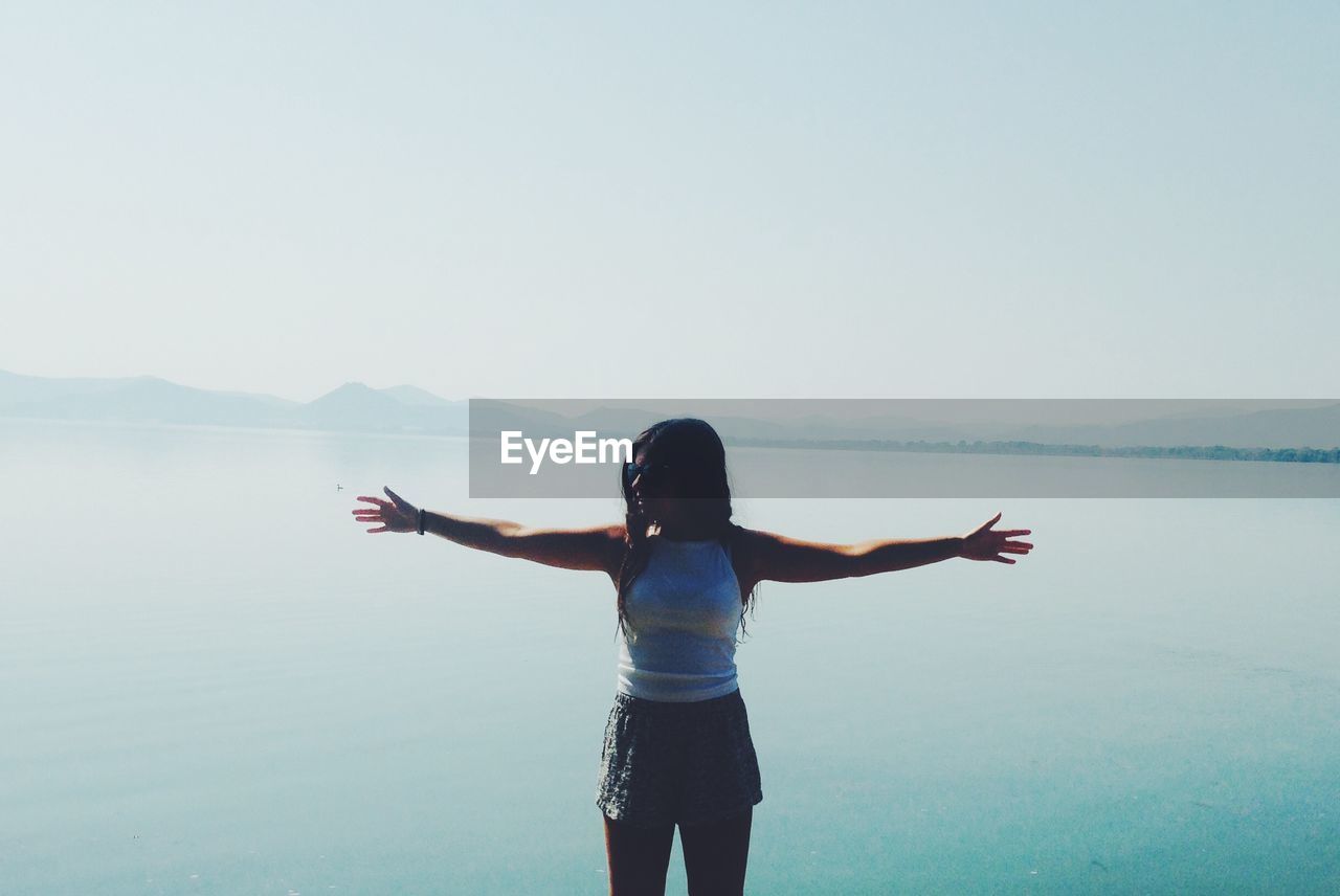 Woman with arms outstretched standing by lake against clear sky