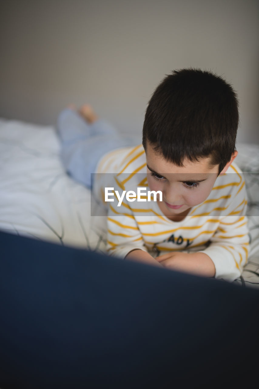 Focused little boy in casual wear browsing laptop while spending free time in bedroom at home