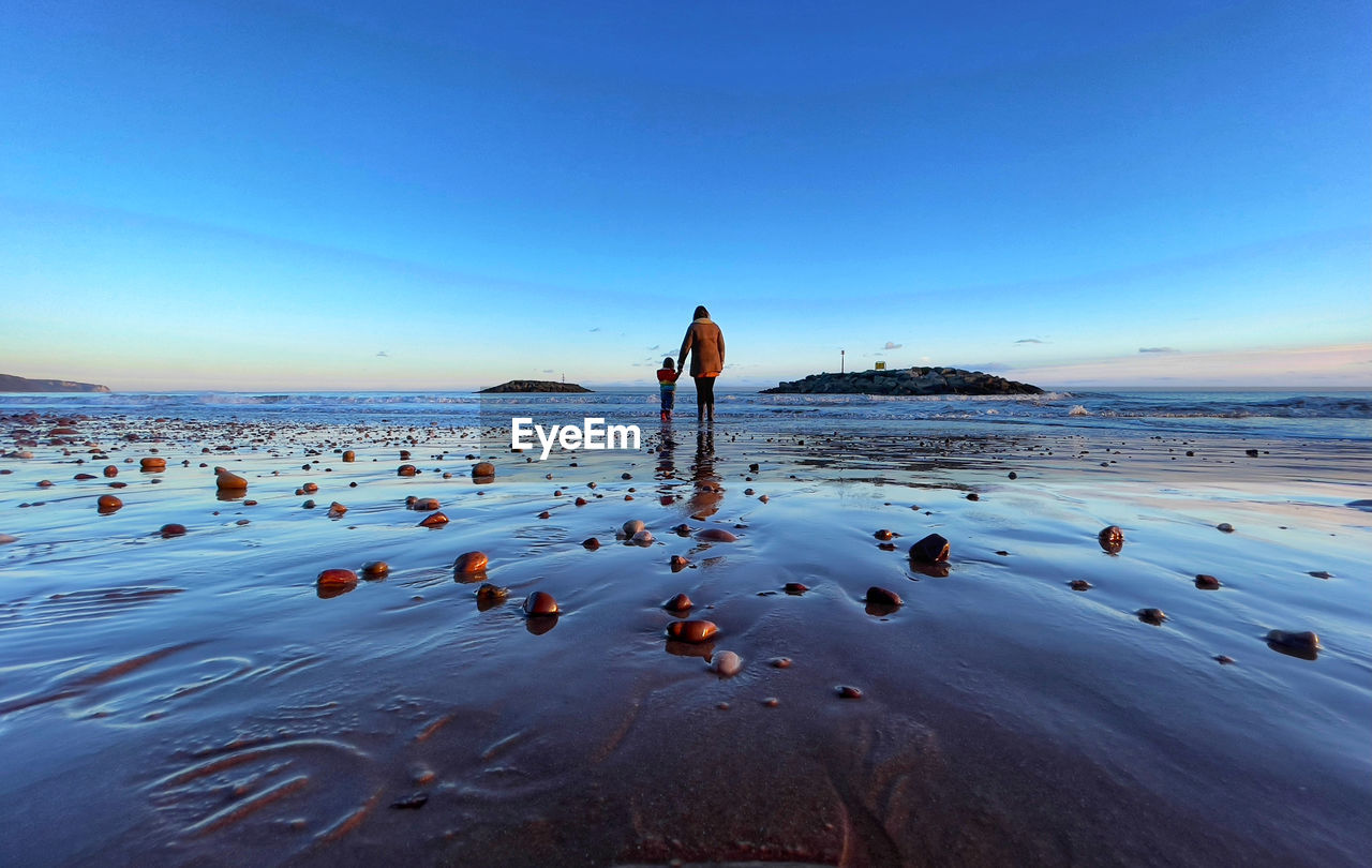 REAR VIEW OF PERSON STANDING AT BEACH AGAINST CLEAR SKY