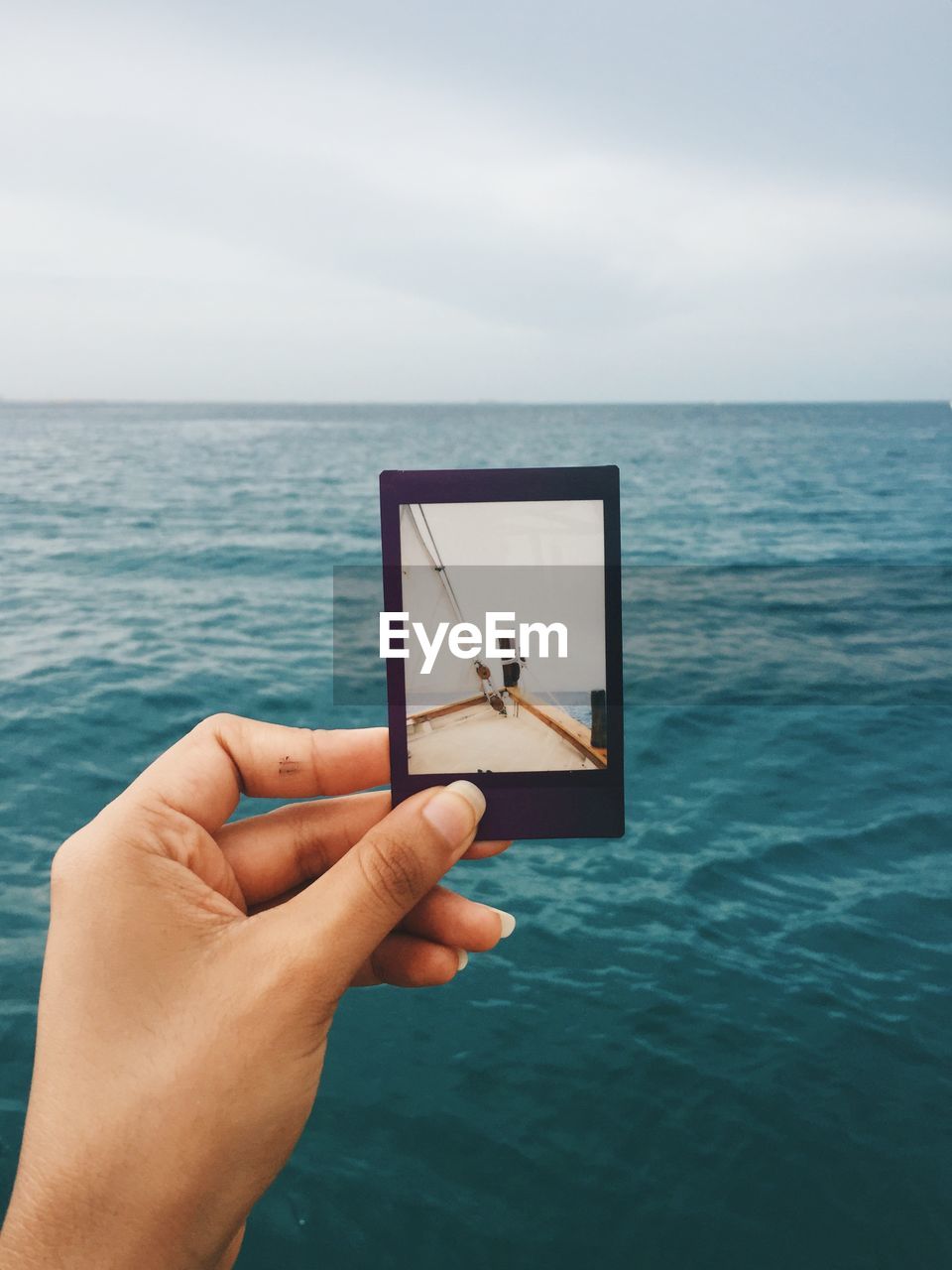 Cropped image of hand holding photograph by sea against sky