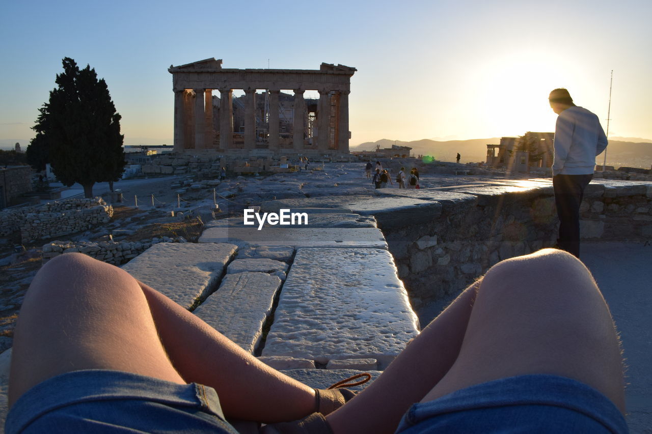 Low section of man with friend visiting parthenon against clear sky during sunset