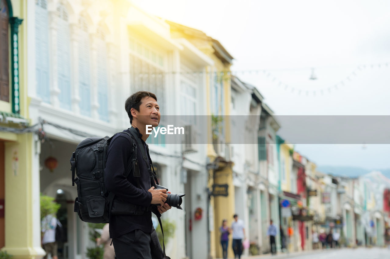 Side view of backpack man with camera standing against buildings in city