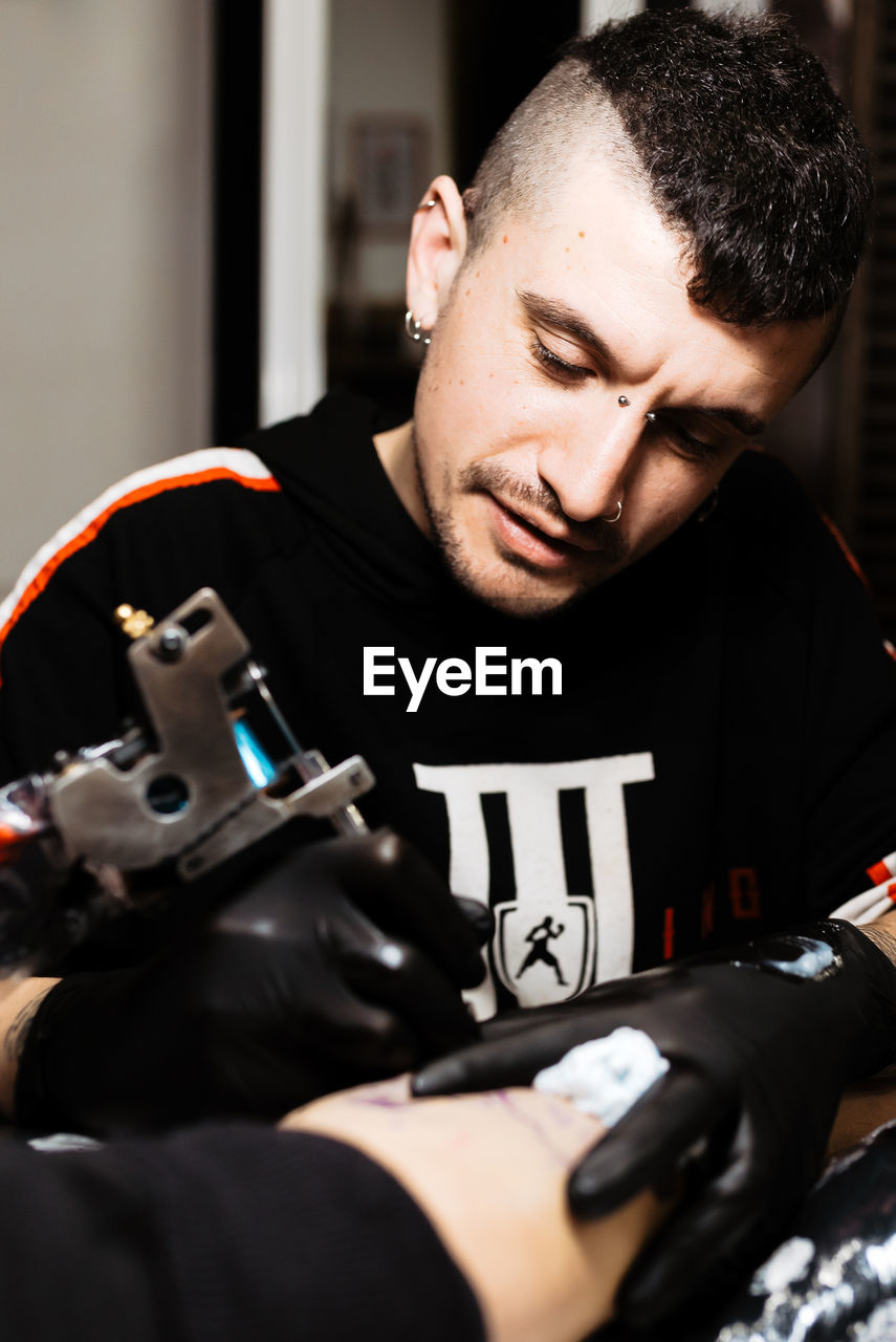 Stylish man with piercing using tattoo machine to make tattoo on leg of crop customer during work in salon