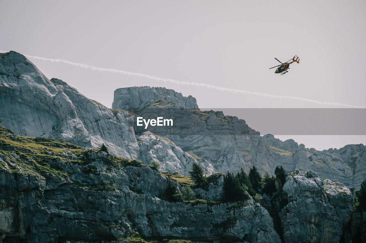 LOW ANGLE VIEW OF HELICOPTER FLYING OVER MOUNTAIN AGAINST SKY