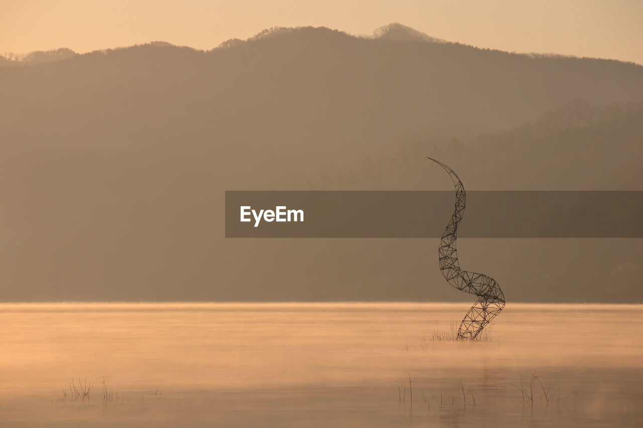 Metal sculpture in lake against silhouette mountain during sunset