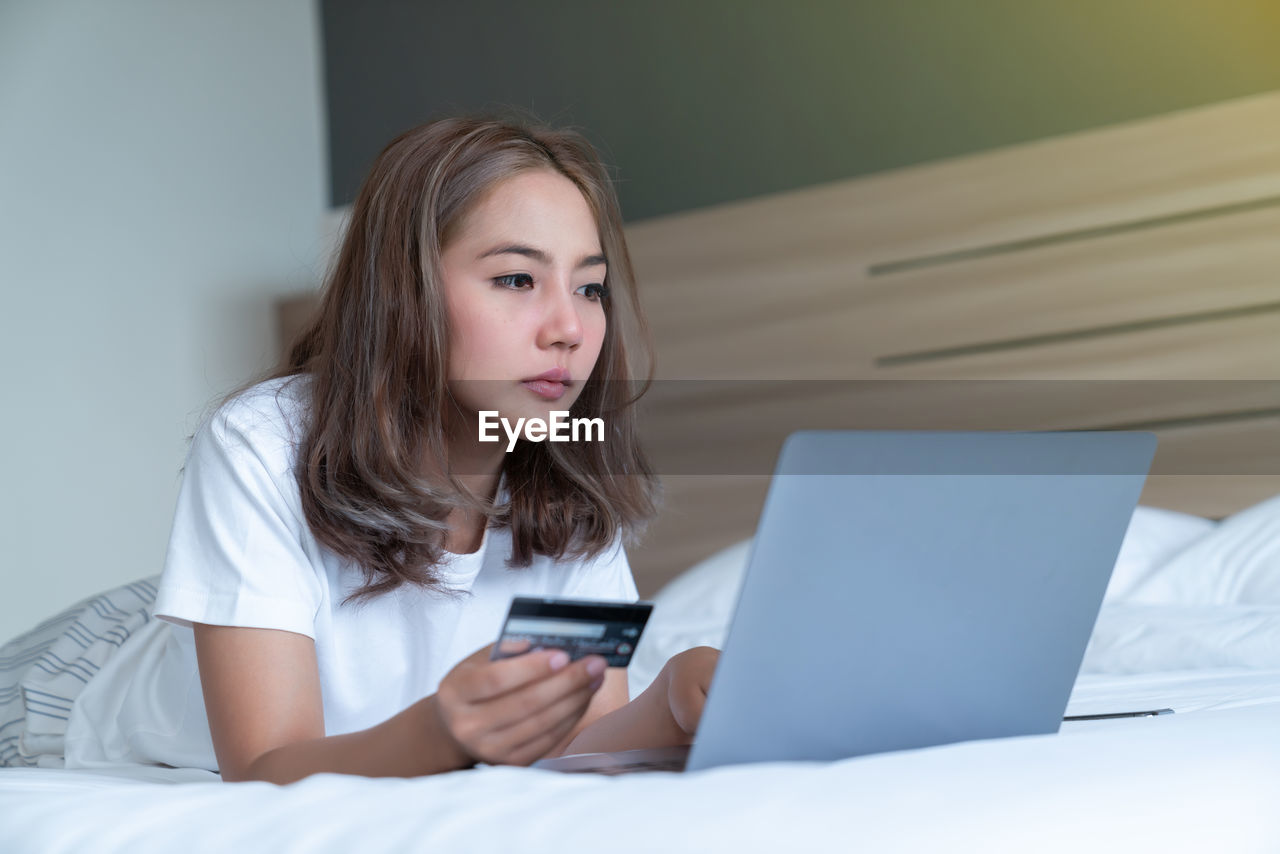 YOUNG WOMAN USING PHONE WHILE SITTING ON BED AT HOME