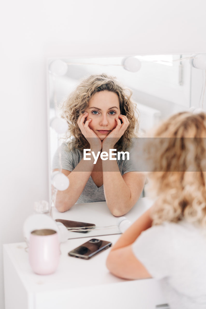Unemotional attractive female in casual shirt touching face and looking at reflection in mirror attentively while sitting at vanity table in bedroom