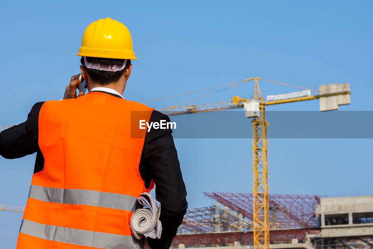 Rear view of engineer using phone at construction site against clear sky