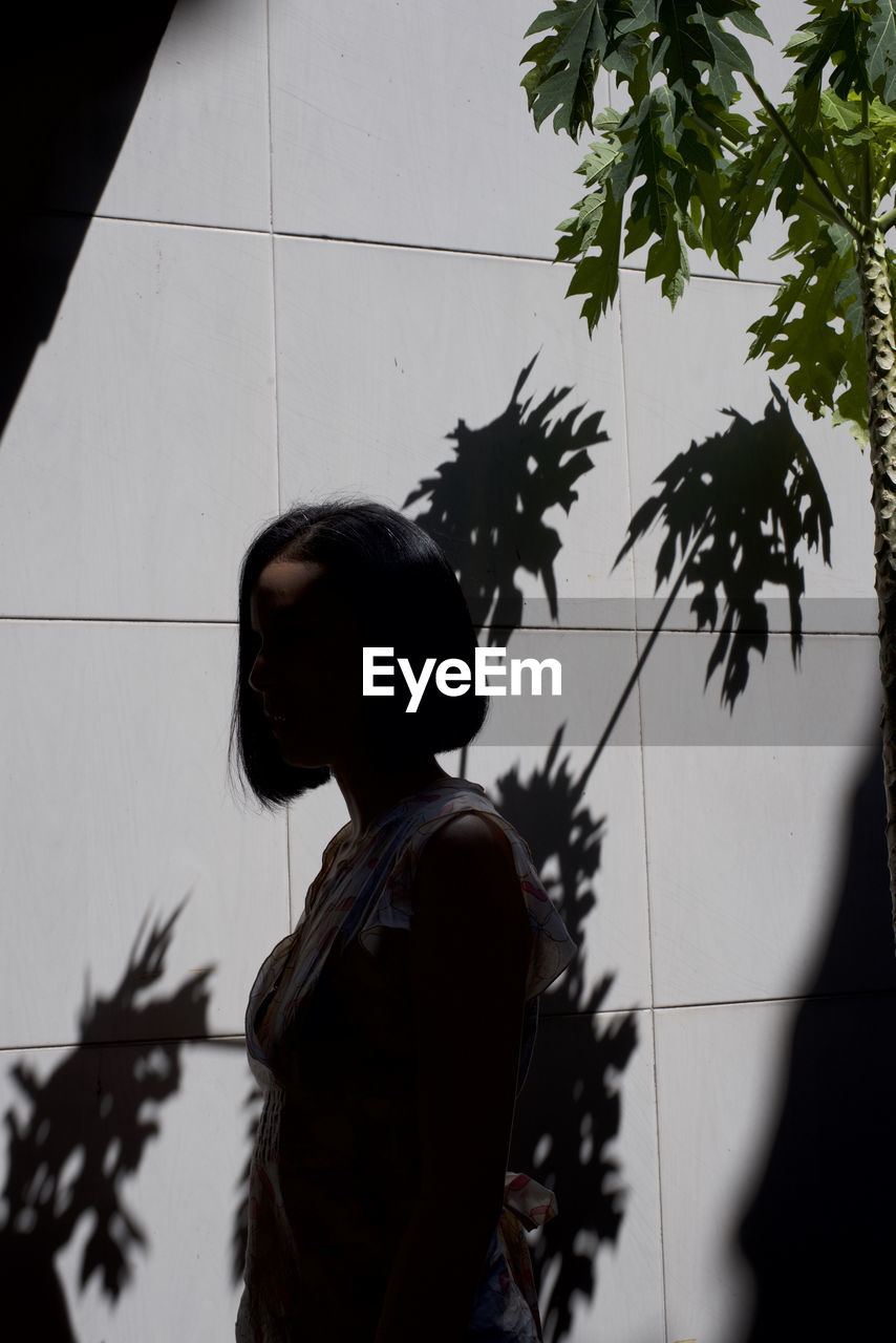 Side view woman standing in tree shadow against wall