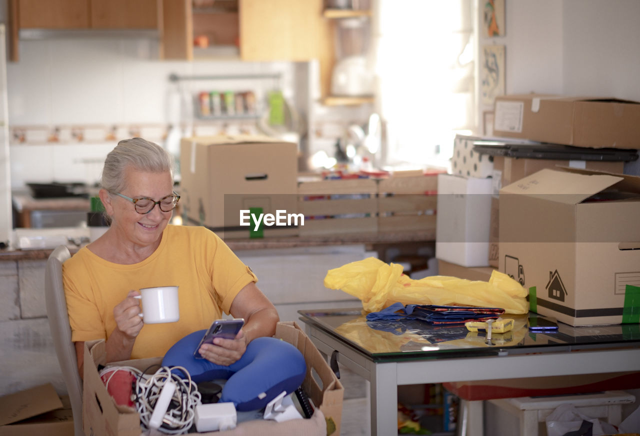 Senior woman drinking coffee cup while using mobile at home