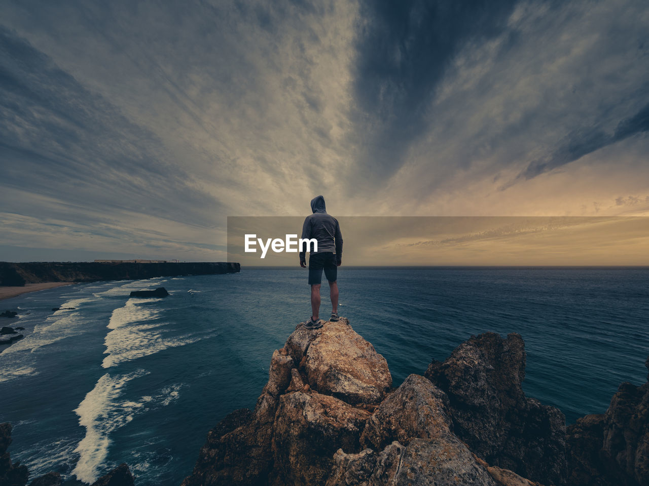 REAR VIEW OF MAN STANDING ON ROCK BY SEA