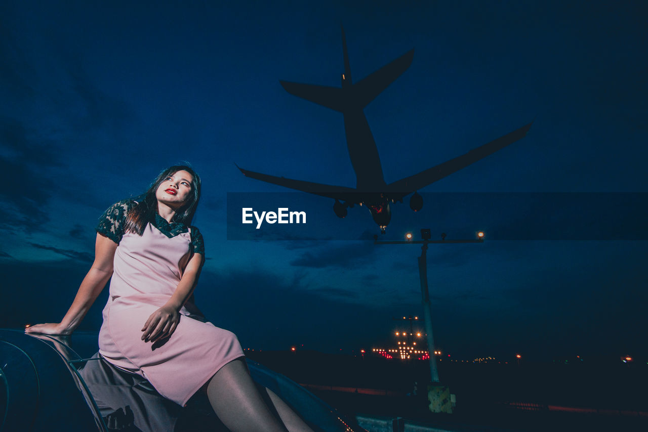 Young woman sitting outdoors while airplane flying against sky at night