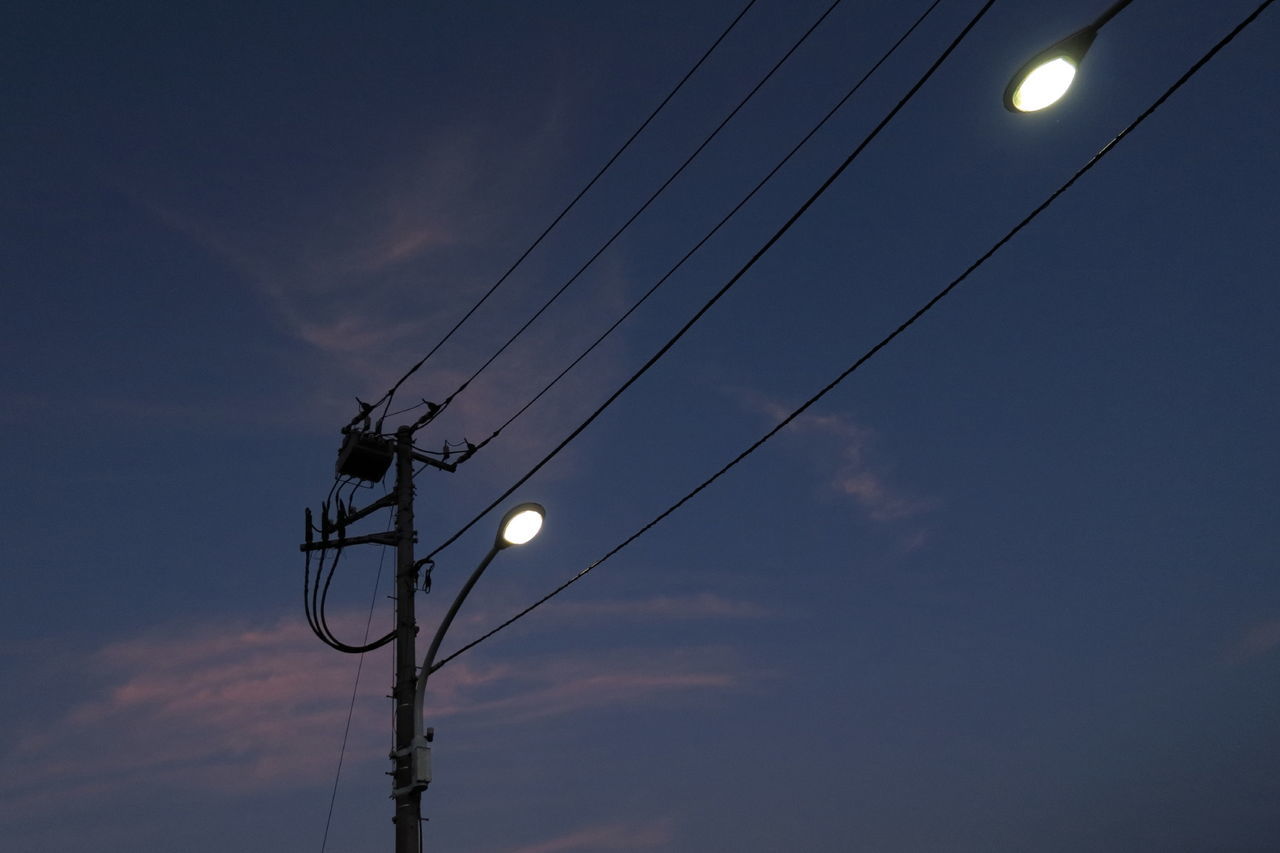 Low angle view of illuminated street lights and electricity pylons against sky at dusk