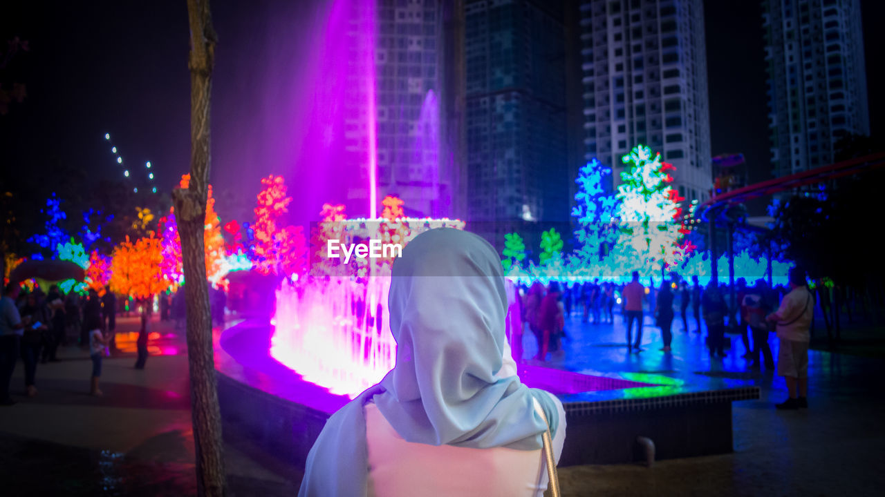 Rear view of woman standing against illuminated fountain at night