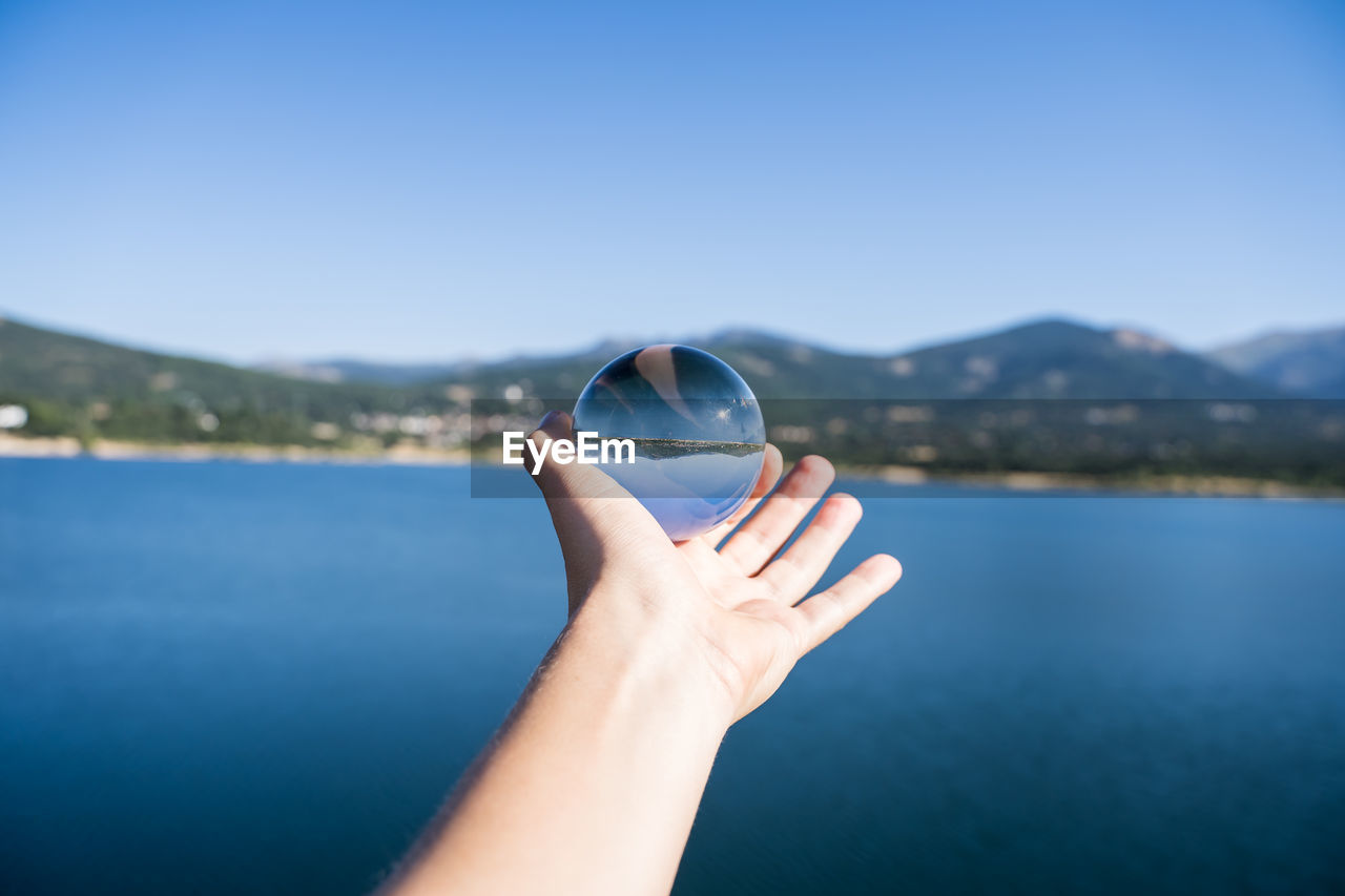Cropped image of hand holding crystal ball against lake