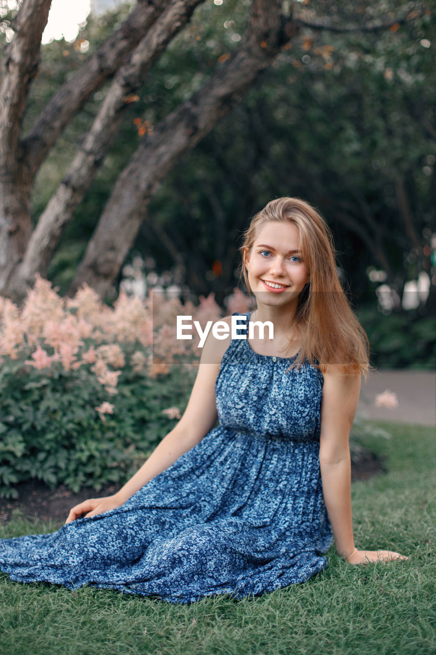 Portrait of smiling young woman sitting on grass against trees