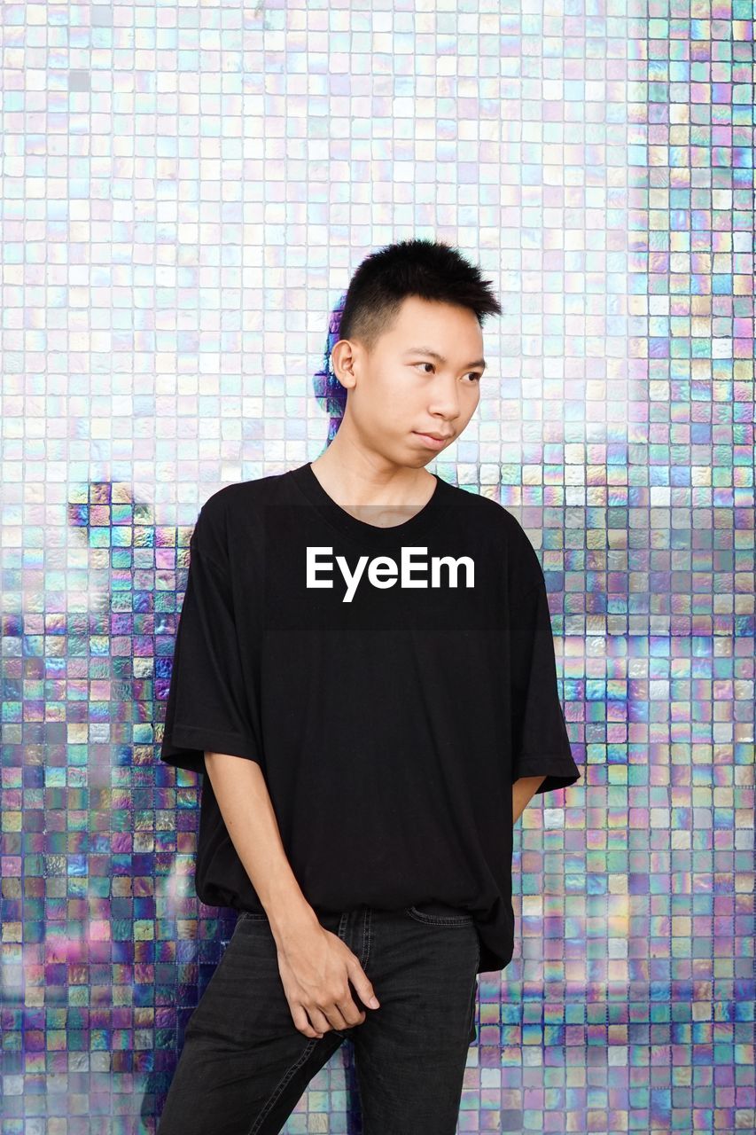 Young man looking away while standing against wall