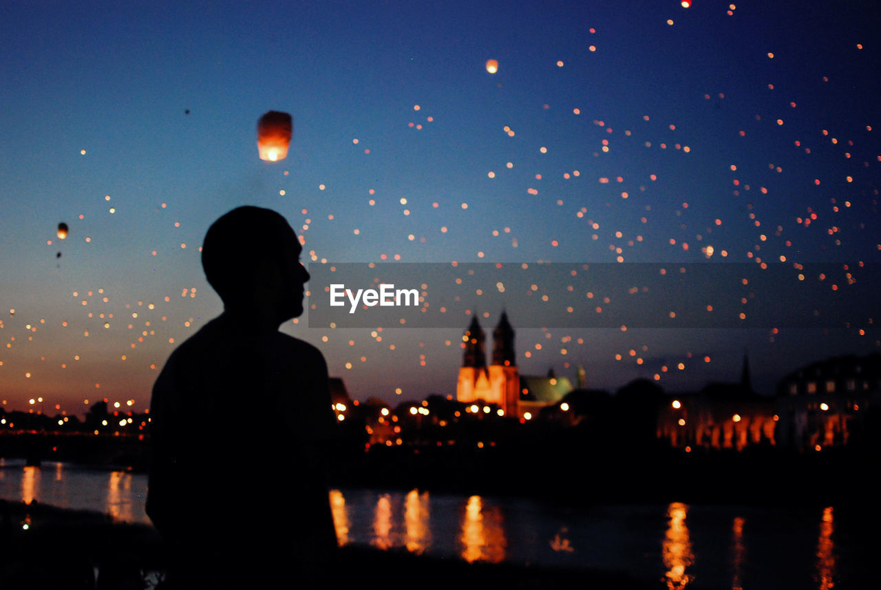 Man looking at sky lanterns flying over river during dusk