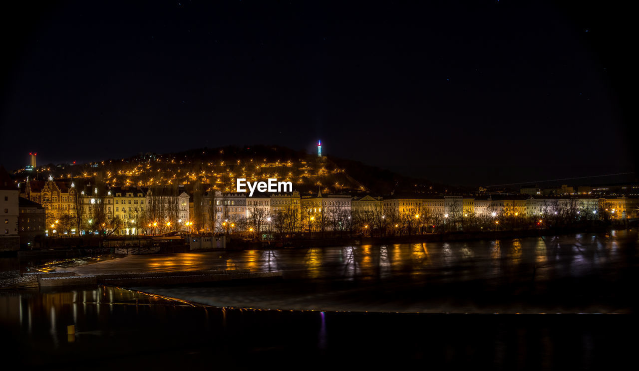 Illuminated buildings by river against sky at night