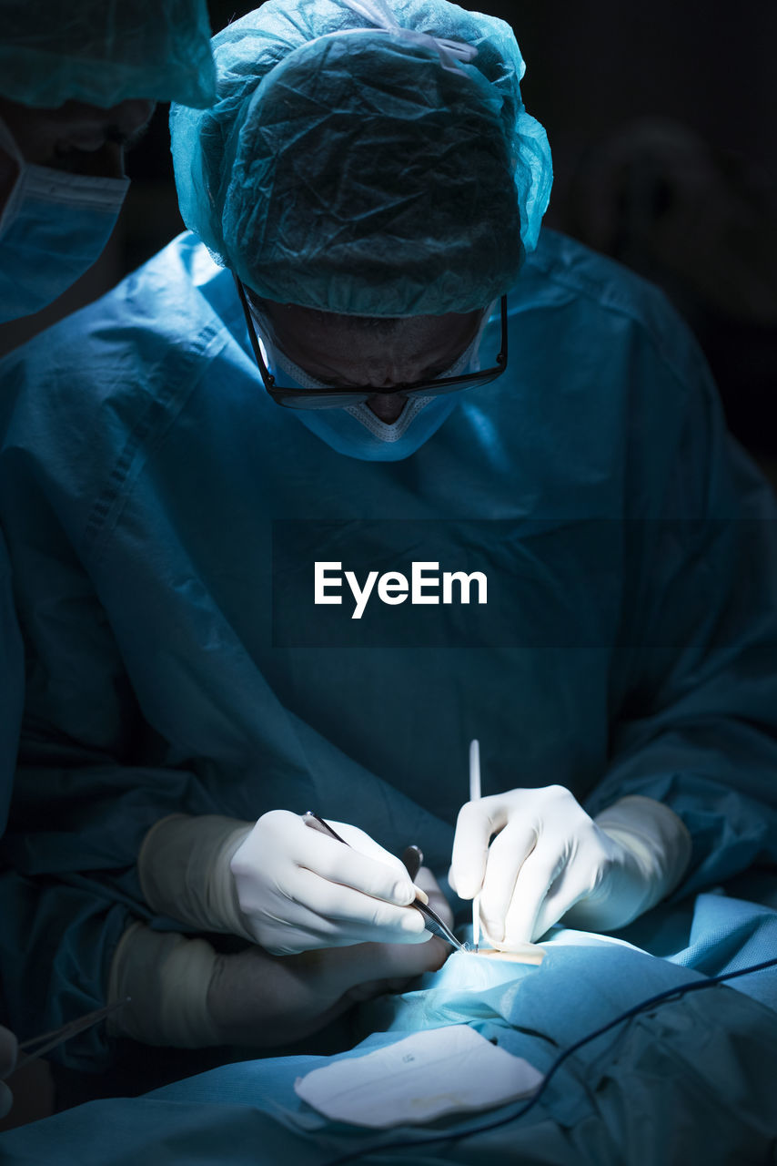 Man in surgeon uniform and his assistant performing surgery in dark operating theater