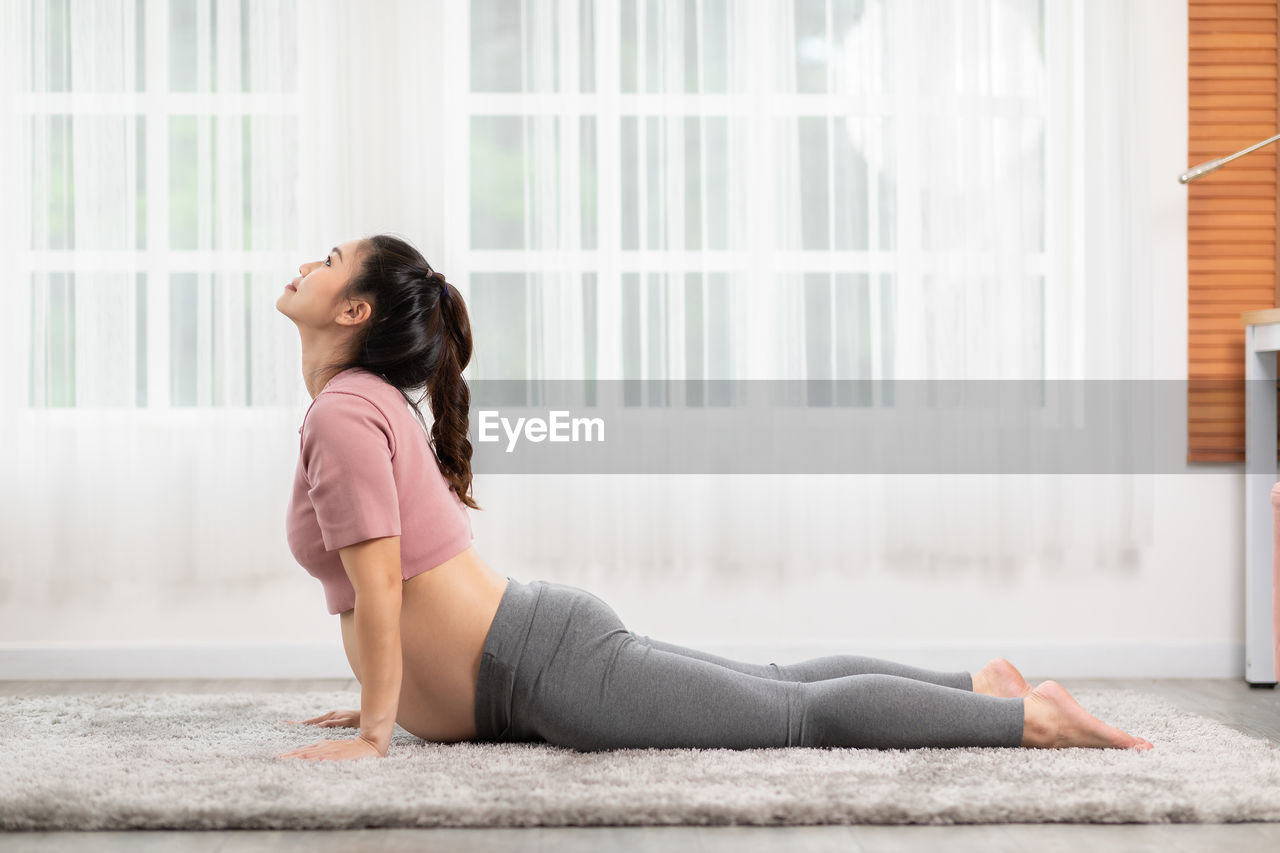 SIDE VIEW OF YOUNG WOMAN SITTING ON WINDOW