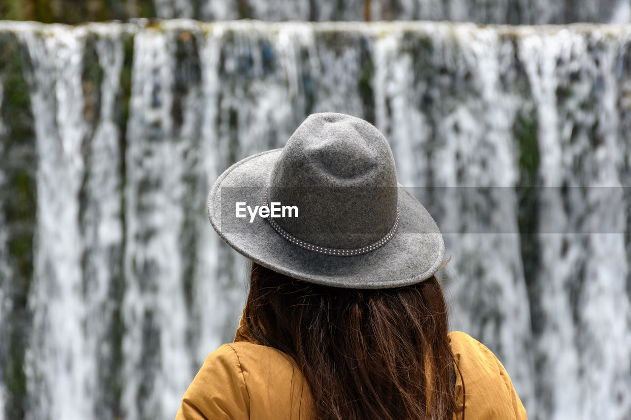 one person, hat, rear view, waterfall, clothing, headshot, adult, women, long hair, spring, sun hat, nature, lifestyles, leisure activity, winter, portrait, water, day, hairstyle, focus on foreground, fashion accessory, fedora, outdoors, young adult, rock, casual clothing, beauty in nature, standing