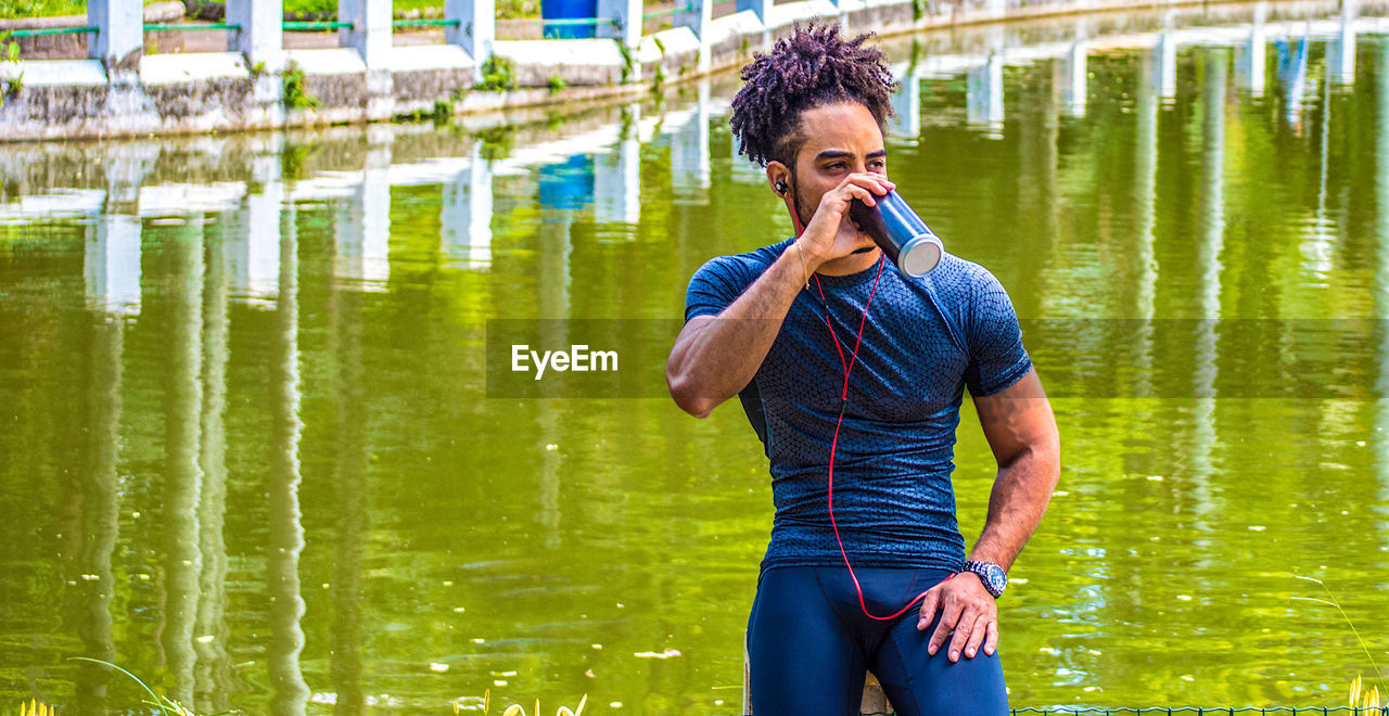 Young man drinking water while listening music against lake