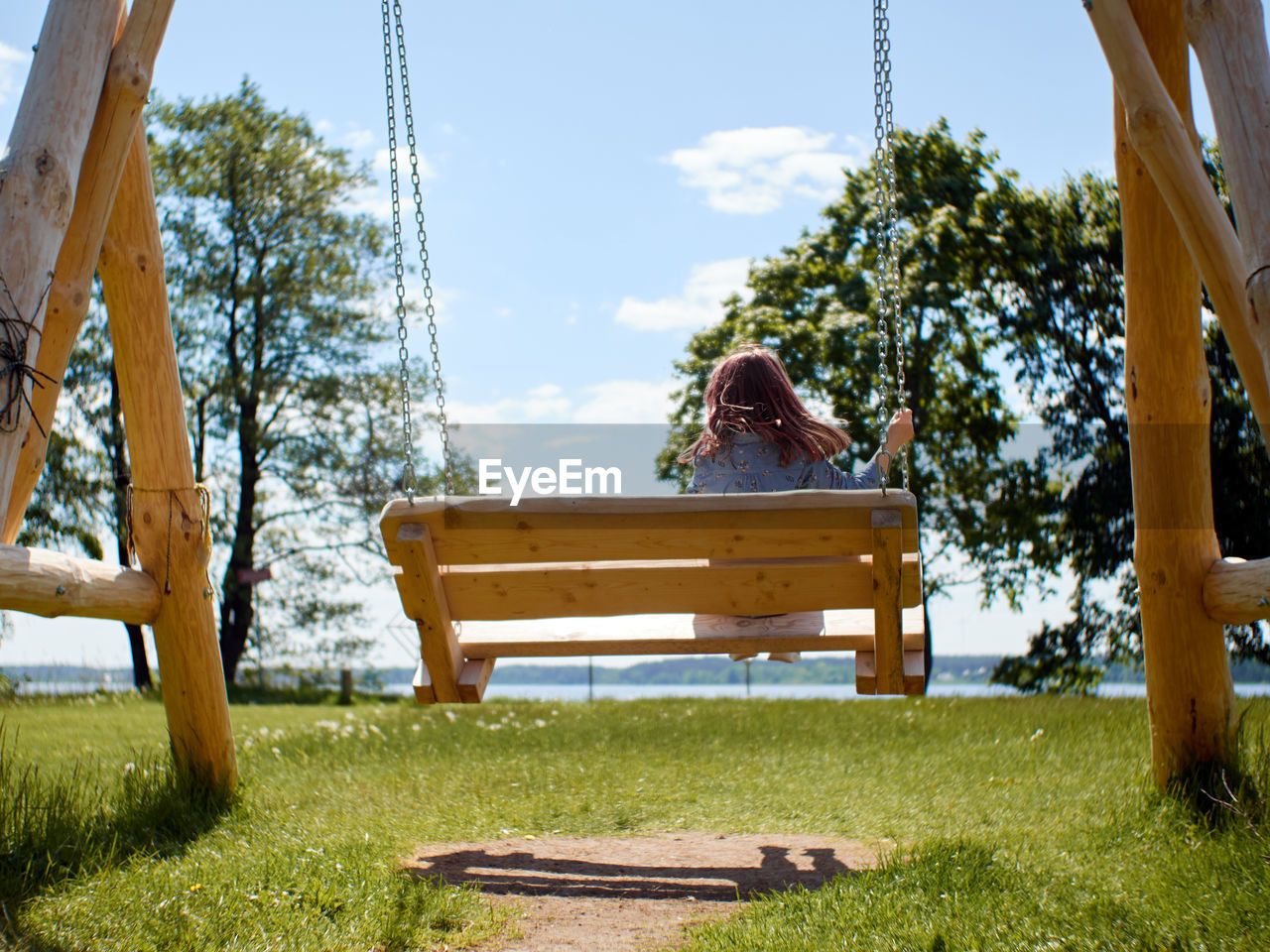 View from back to kid with red hairs riding on a wooden swing on the lawn against blue sky.