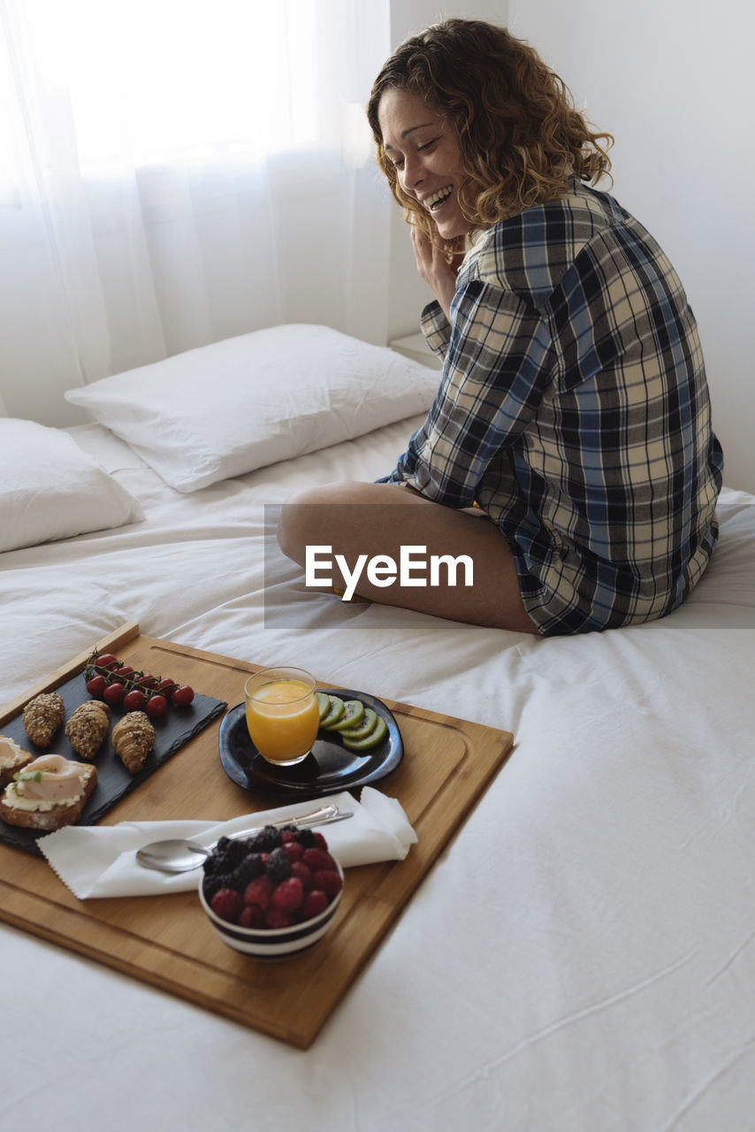 Woman with curly hair and plaid shirt having a breakfast on bed.