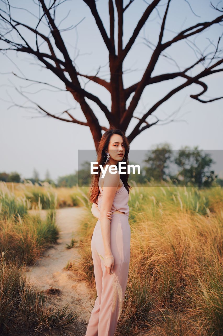 Portrait of young woman standing on grassy land against bare tree
