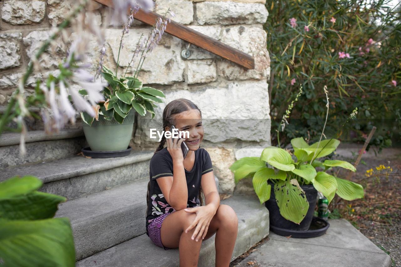 Girl on phone amidst garden plants, enjoying a peaceful rural holiday.