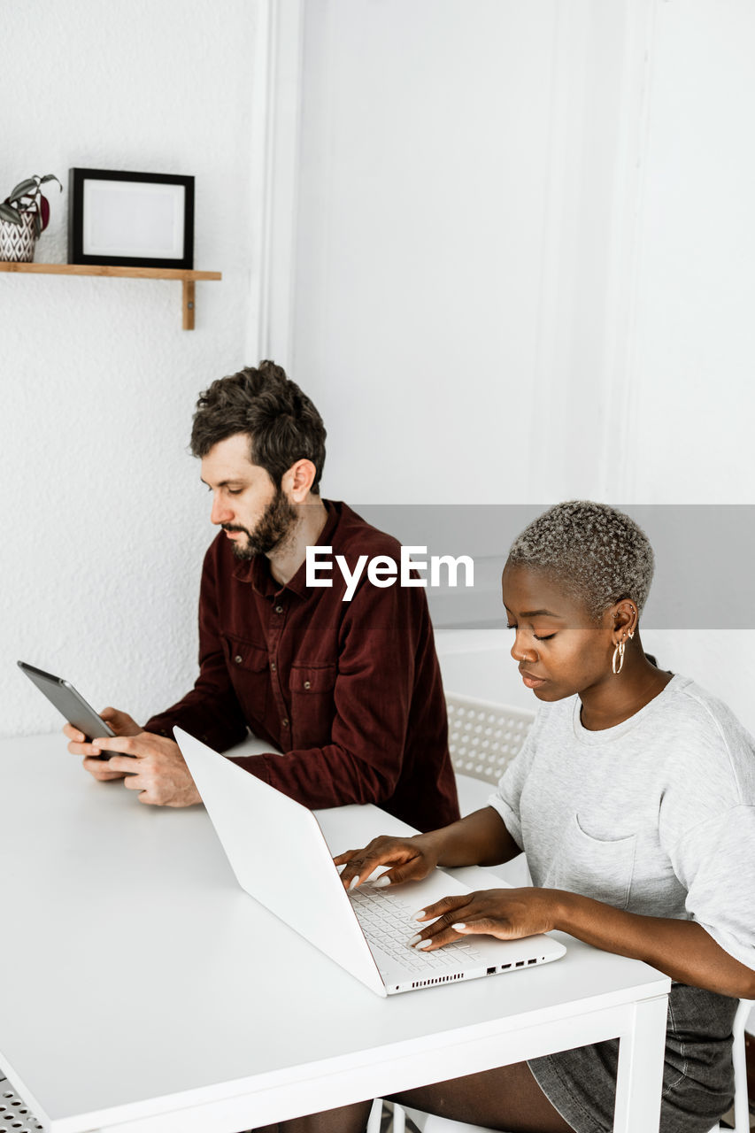 Side view from above of contemporary african american woman sitting at table and working with laptop with young man surfing digital table at home