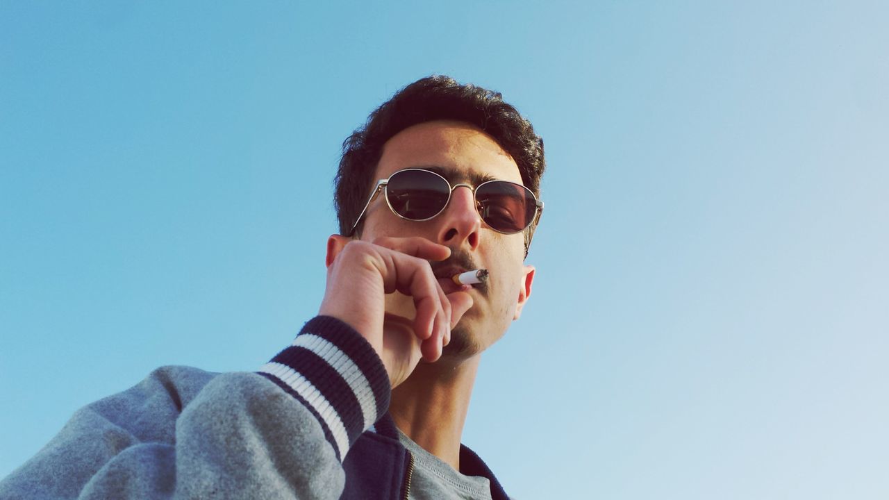 Low angle portrait of man wearing sunglasses while smoking against clear blue sky