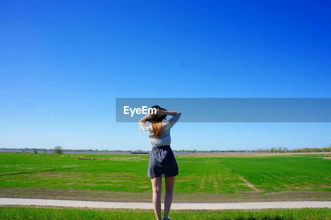 Rear view of woman wearing hat standing on grassy field against sky during sunny day