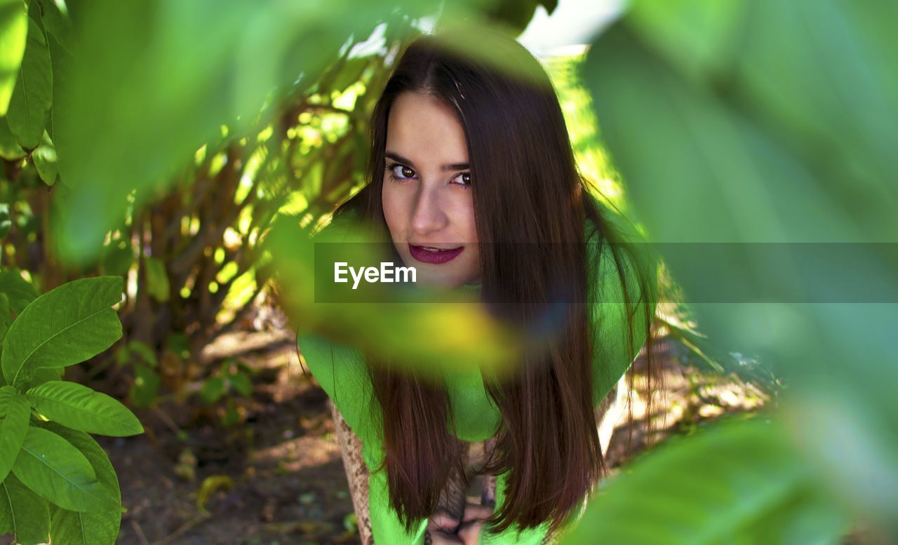 Portrait of young woman seen through leaves standing against plants