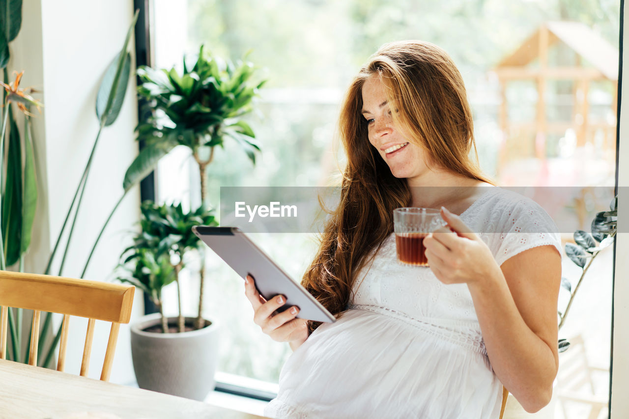 Charming pregnant woman drinks a drink and looks at the screen of a computer tablet.