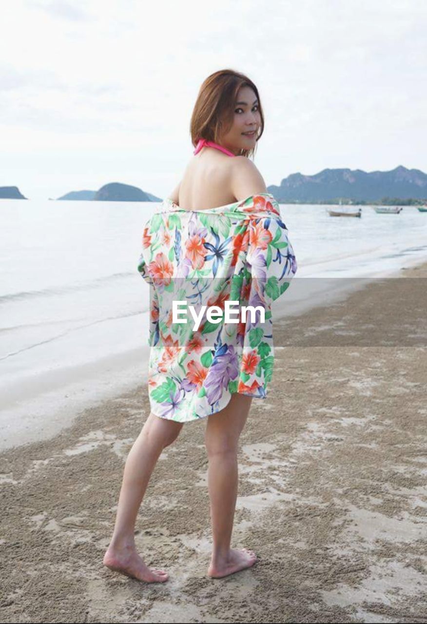 Portrait of young woman standing at beach against sky