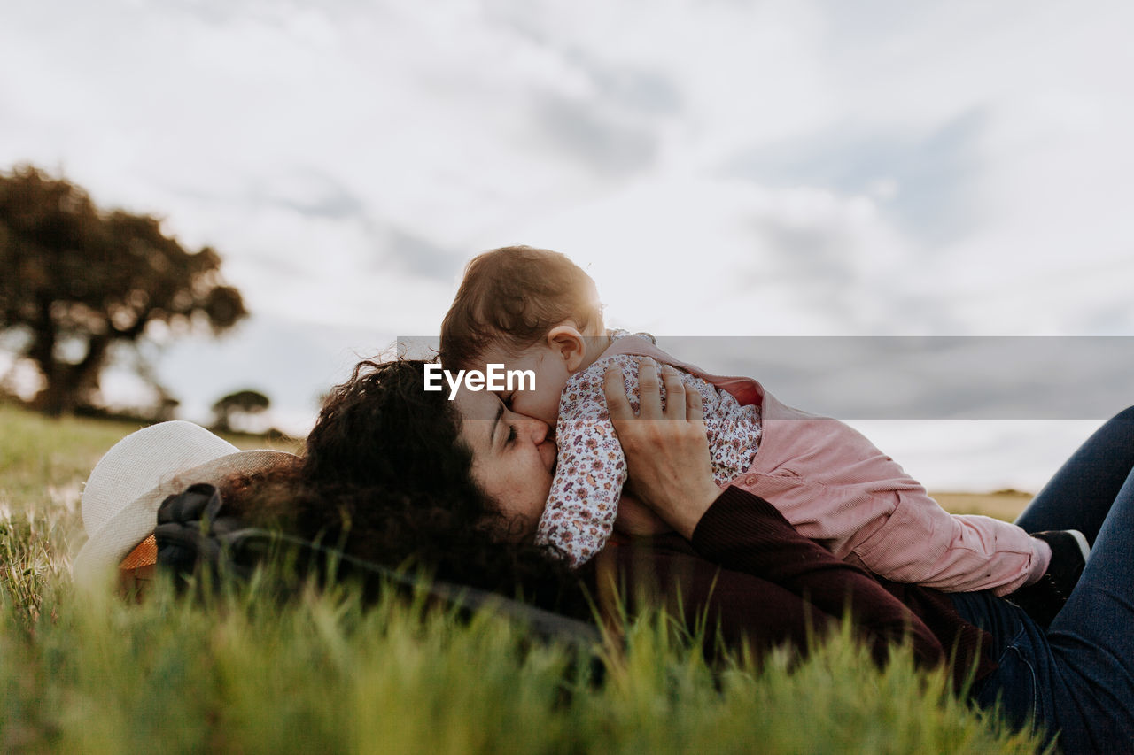 Mother kissing daughter while lying on lawn against sky